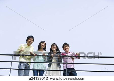 Stock Image - Four young people leaning on the railing, side by side, low angle view, blue background, copy space, Japan. Fotosearch