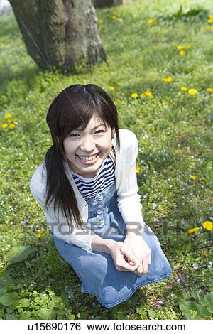 Stock Photograph - Portrait of a woman looking at camera and crouching down on lawn, high angle view, Japan. Fotosearch