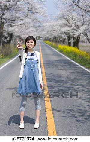 Picture - Portrait of a woman making peace sign on the road lined with cherry trees, smiling and looking at camera, front view, Japan. Fotosearch