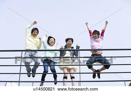 Stock Image - Portrait of four young people jumping behind the railing, low angle view, blue background, Japan. Fotosearch