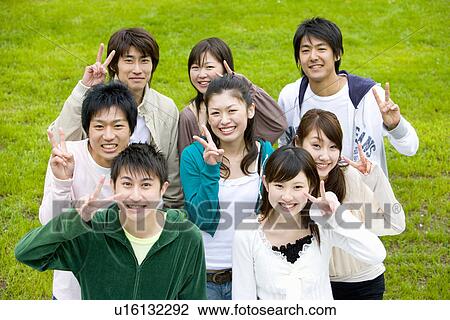 Stock Image - Portrait of medium group of young people making peace sign on lawn, smiling and looking at camera, high angle view, Japan. Fotosearch