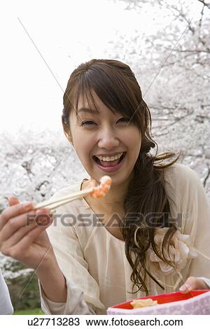 Stock Image - Portrait of woman holding shrimp with chopsticks, smiling and looking at camera, front view, Japan. Fotosearch