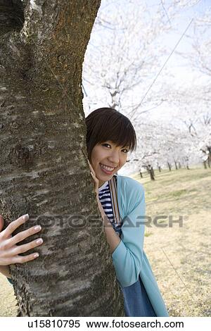Stock Photography - Portrait of woman standing behind the cherry tree, smiling and looking at camera, front view, Japan. Fotosearch