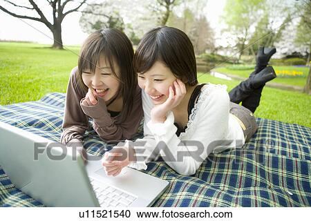 Stock Image - Two young women looking at laptop, lying on sheet, front view, Japan. Fotosearch