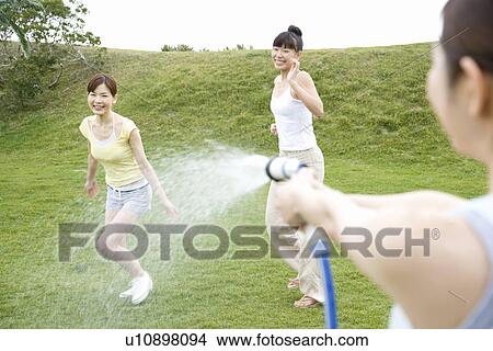 Young woman spraying water to friends, side view, rear view View Large Photo Image Picture - Young woman spraying water to friends, side view, rear view. Fotosearch