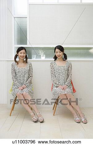 Stock Photograph - Portrait of twin sisters sitting on chairs, smiling and looking at camera, front view. Fotosearch