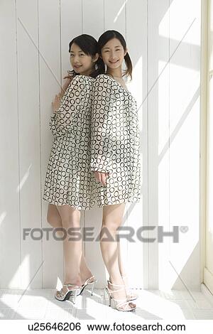 Stock Photograph - Portrait of twin sisters standing back to back, smiling and looking at camera, side view. Fotosearch