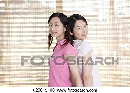 Stock Image - Portrait of Two Young Twin Sisters, Back to Back, Smiling. Side View. Fotosearch