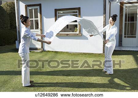 Stock Photograph - Twin sisters holding a sheet on garden, smiling, side view. Fotosearch