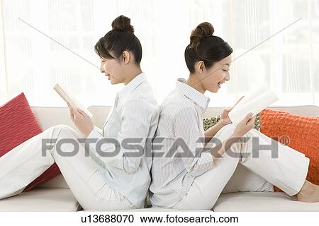 Twin sisters sitting on couch and reading books, back to back, smiling, side view View Large Photo Image Stock Image - Twin sisters sitting on couch and reading books, back to back, smiling, side view. Fotosearch