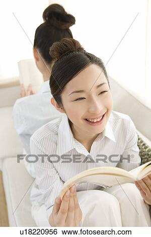 Stock Photograph - Twin sisters sitting on couch and reading books, back to back, smiling, high angle view, rear view, front view. Fotosearch