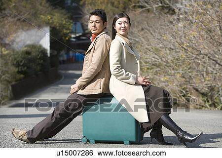 Stock Photograph - Japanese couple on the road, sitting on a suitcase, side view, Japan. Fotosearch