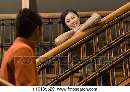 Stock Photo - Woman leaning on the stair rail and smiling to man, front view, side view, Japan. Fotosearch