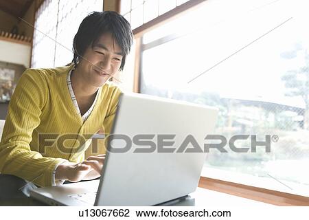 Stock Image - Young man smiling and using laptop, front view, Japan. Fotosearch