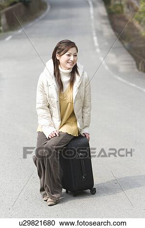 Stock Image - Young woman sitting on a suitcase on the road, front view, Japan. Fotosearch