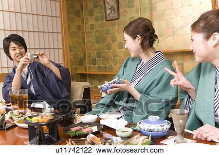 Stock Image - Three young people having dinner, man taking a picture, front view, side view, Japan. Fotosearch
