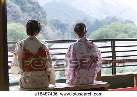 Stock Photograph - Two women in Yukata sitting on bench and looking outside, rear view, Japan. Fotosearch
