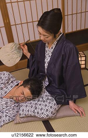 Stock Image - Woman fanning man, man resting his head on her lap in the room, front view, high angle view, Japan. Fotosearch
