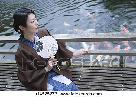 Stock Image - Woman in Yukata holding a Japanese fan and looking at the carp in pond, side view, Japan. Fotosearch