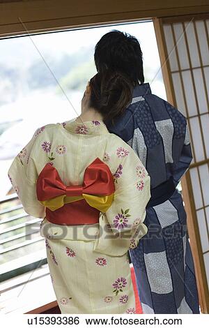 Stock Photograph - Young couple in Yukata looking outside at the window, woman leaning on man, rear view, Japan. Fotosearch