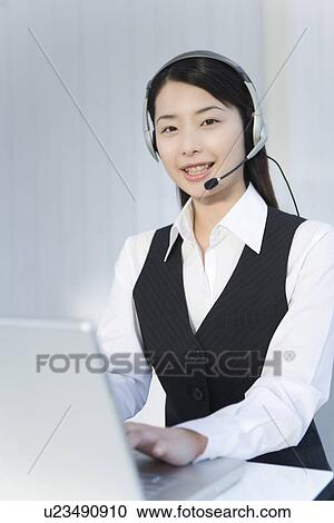 Stock Image - Portrait of a woman wearing hands-free headset and using laptop, looking at camera, front view. Fotosearch