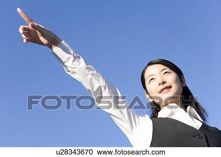Stock Image - Woman pointing forward under the blue sky, low angle view, blue background. Fotosearch