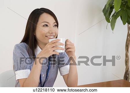 Stock Image - Young woman drinking coffee in office. Fotosearch