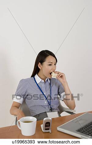Stock Image - Young woman eating sandwiches at desk. Fotosearch