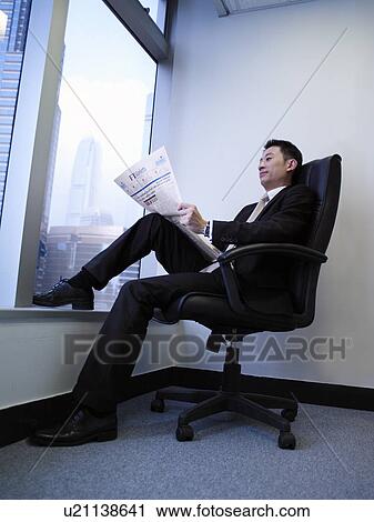 Stock Image - Businessman sitting in chair reading newspaper in front of window. Fotosearch