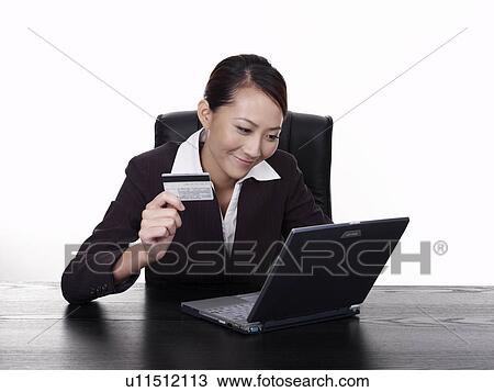 Stock Image - Businesswoman using laptop holding credit card, studio shot. Fotosearch