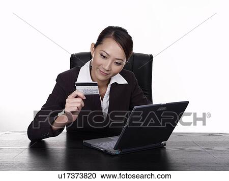 Stock Image - Businesswoman using laptop holding credit card, studio shot. Fotosearch