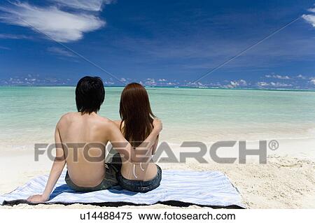 Stock Photography - Young couple sitting on beach, man putting arm around woman's shoulders, Saipan, USA . Fotosearch