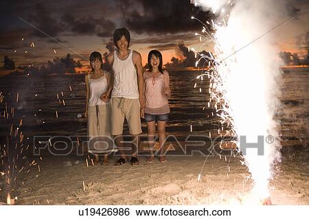 Stock Photograph - Three young people looking at lighting firework on beach, long exposure, Saipan, USA . Fotosearch