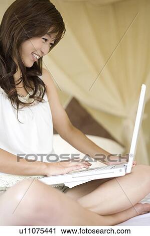 Stock Image - Young woman sitting on a bed using laptop, Saipan. Fotosearch