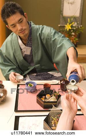 Stock Image - Japanese couple in Yukata having dinner, man pouring Sake, front view, Japan. Fotosearch