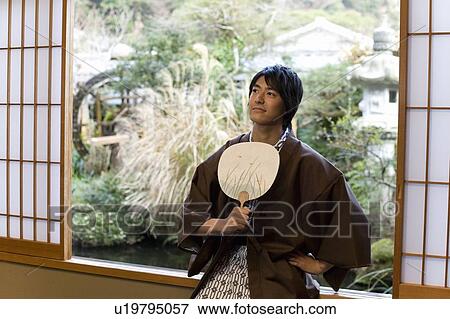 Stock Photo - Man in Yukata holding Japanese fan and sitting at the window, front view, Japan. Fotosearch