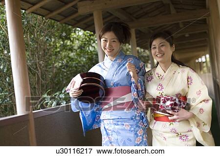Stock Photo - Portrait of two women in Yukata holding wooden tubs, smiling and looking at camera, front view, Japan. Fotosearch