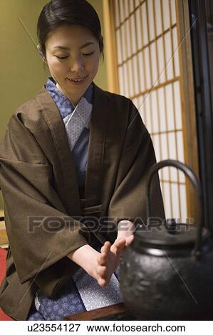 Stock Photo - Woman in Yukata sitting in front of a tea kettle in room, front view, Japan. Fotosearch