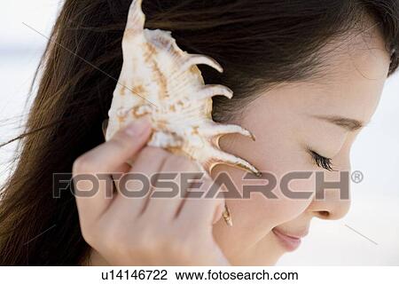 Stock Image - Young woman listening to seashell. Fotosearch