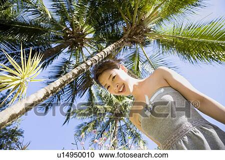 Stock Image - Young woman standing by palm tree. Fotosearch