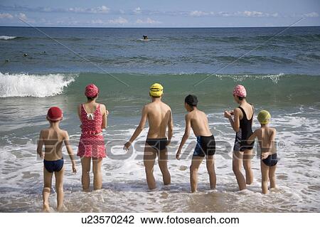 Stock Image - Six children standing in water and watching the surf. Fotosearch