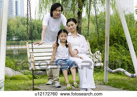 Stock Image - Young family with daughter sitting on the glider and looking at the camera. Fotosearch