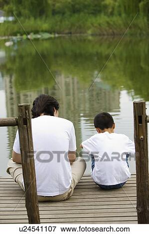 Stock Photo - Young father with son sitting on the wooden board near the lakeside. Fotosearch