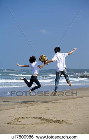 Stock Photograph - Young couple holding a bunch of flowers and jumping on the beach. Fotosearch