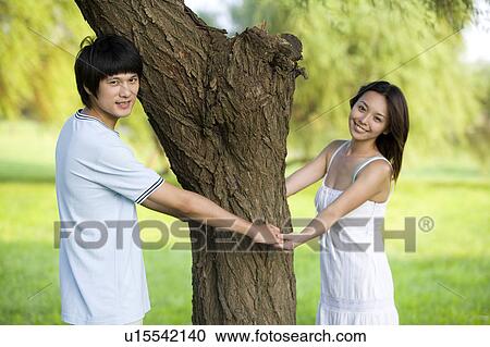 Stock Image - Young couple holding hands around the tree trunk. Fotosearch