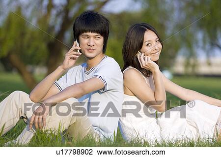 Stock Image - Young couple sitting on the lawn and holding mobile phone, back to back. Fotosearch