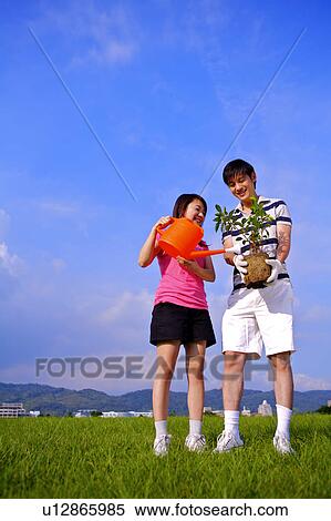 Stock Photography - Young couple standing on the lawn, holding a small tree and watering. Fotosearch