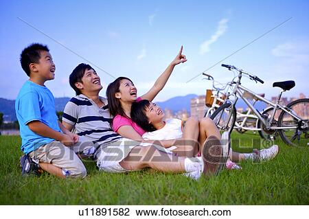 Stock Image - Young family with two children sitting on the lawn together, looking at the sky and smiling happily. Fotosearch