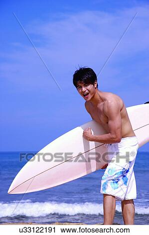 Stock Image - Man holding the surfboard and making a pose on beach. Fotosearch