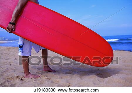 Stock Image - Man holding the surfboard and standing on the beach. Fotosearch